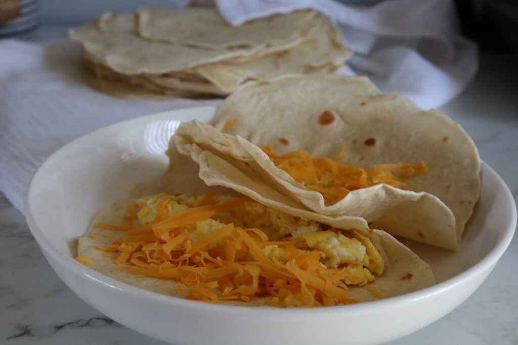close up of two egg and cheese tacos in sourdough tortillas on a plate with stack of tortillas on towel in the background