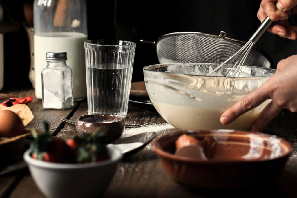 glass of water on a table next to bowl of batter and other items out of focus
